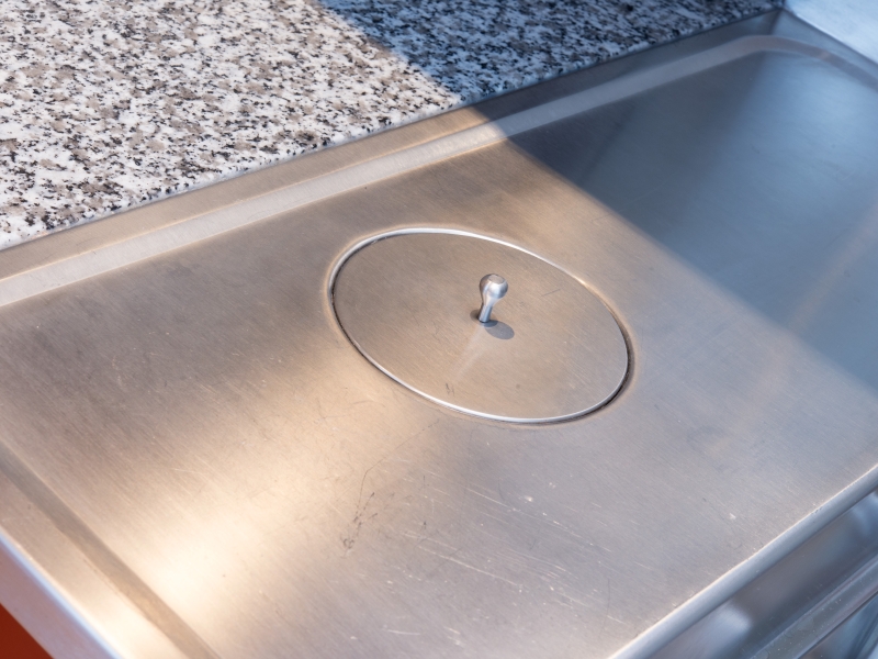 Mit Deckel verschlossener Müll-Abwurfschacht, fugenlos in Edelstahl-Arbeitsplatte eingeschweißt (Arbeitsfläche mit Wulstrand abgesenkt)